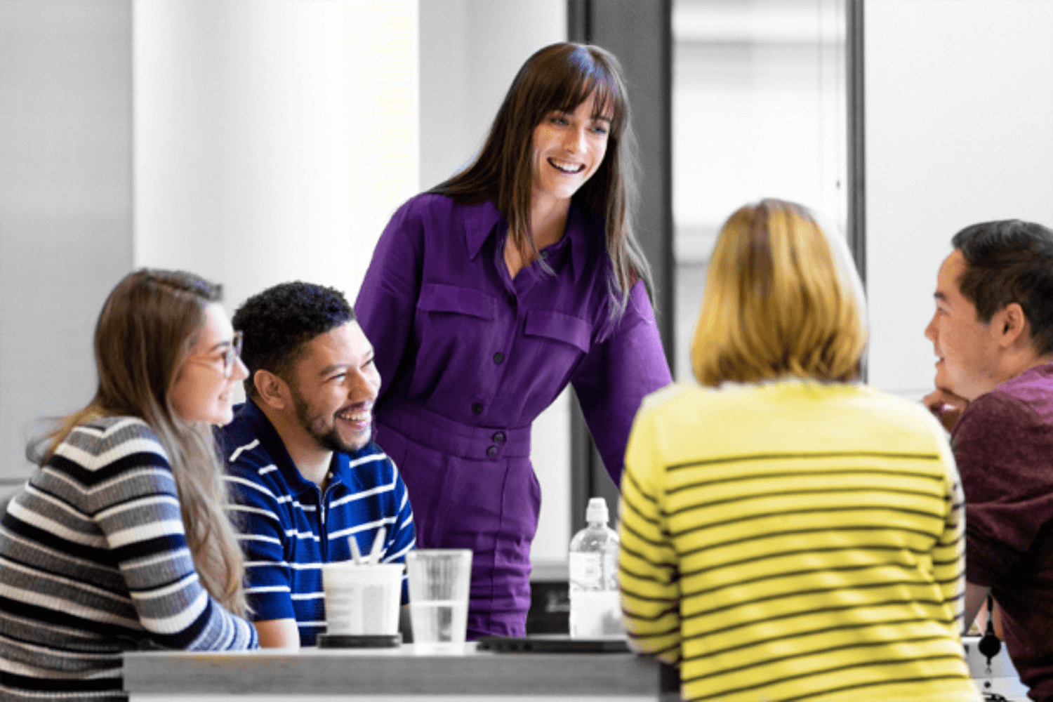 Black and White image, where only clothes and skin tone are in colour. A woman in a purple dress is stood up over a table of four colleagues.