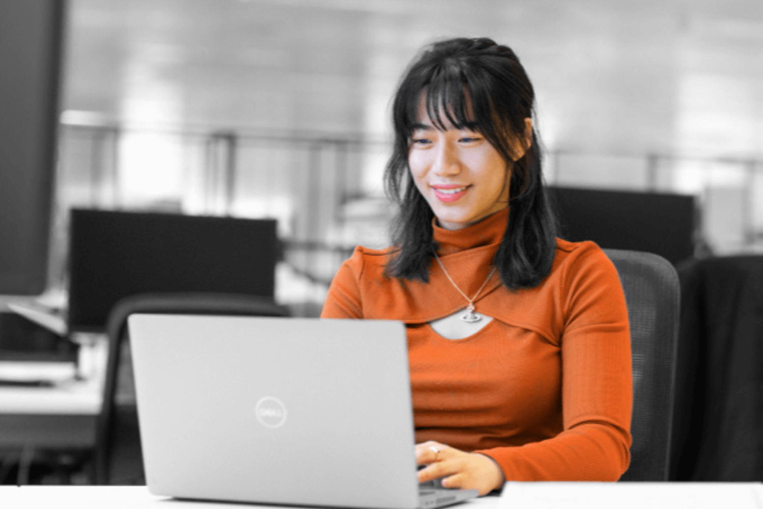 Black and White image, where only clothes and skin tone are in colour. A young woman in an orange top works at a laptop. 