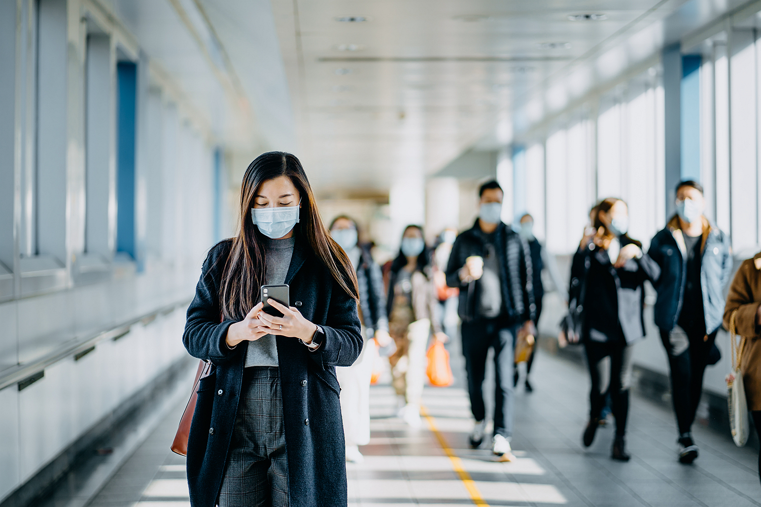 Asian woman with protective face mask using smartphone while commuting in the urban bridge in city against crowd of people.